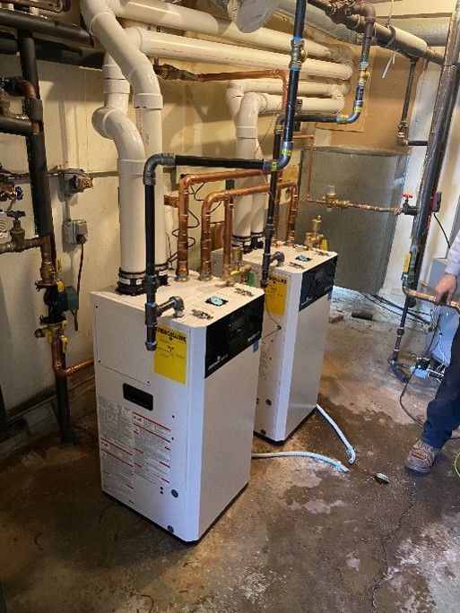 Two white commercial water heaters connected with black iron and copper piping in a basement utility room.