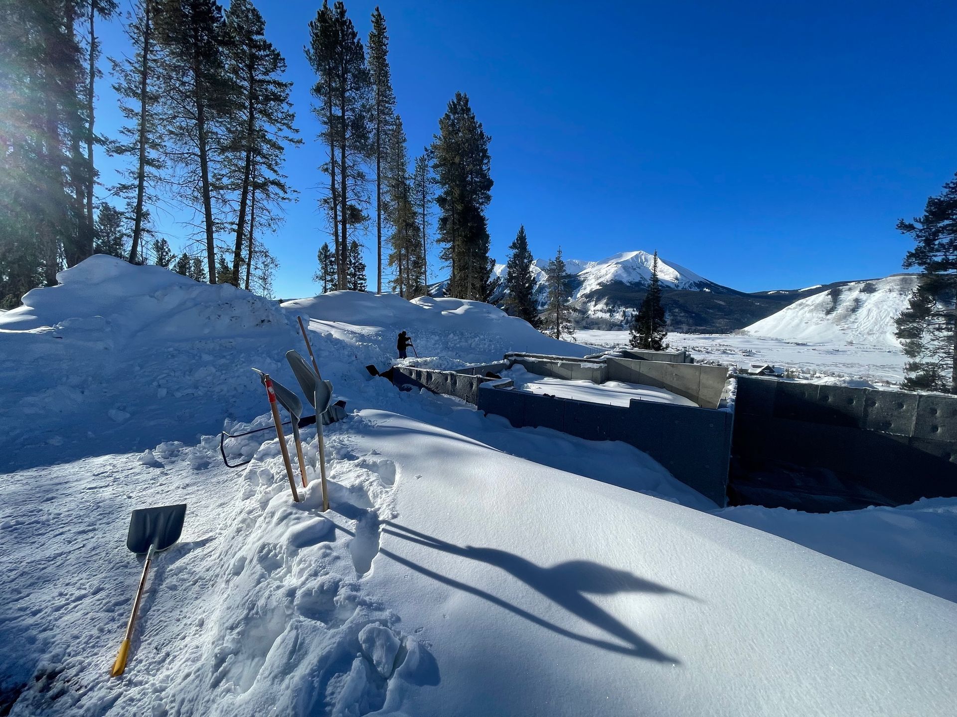 Snowy landscape with shovels and tools in the foreground, mountains and trees in the background under a blue sky.
