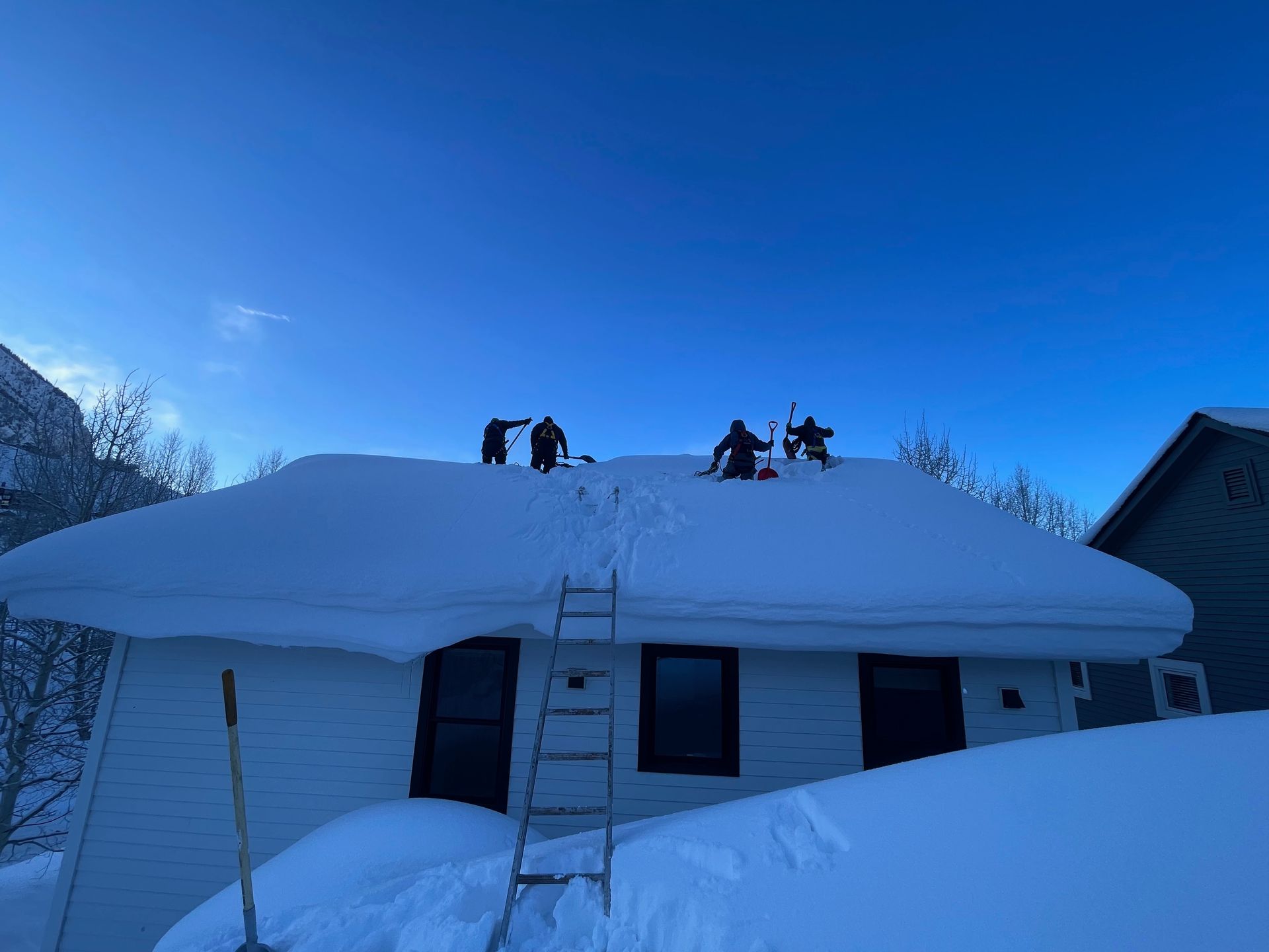People shoveling snow off a white house roof under a clear, blue sky.