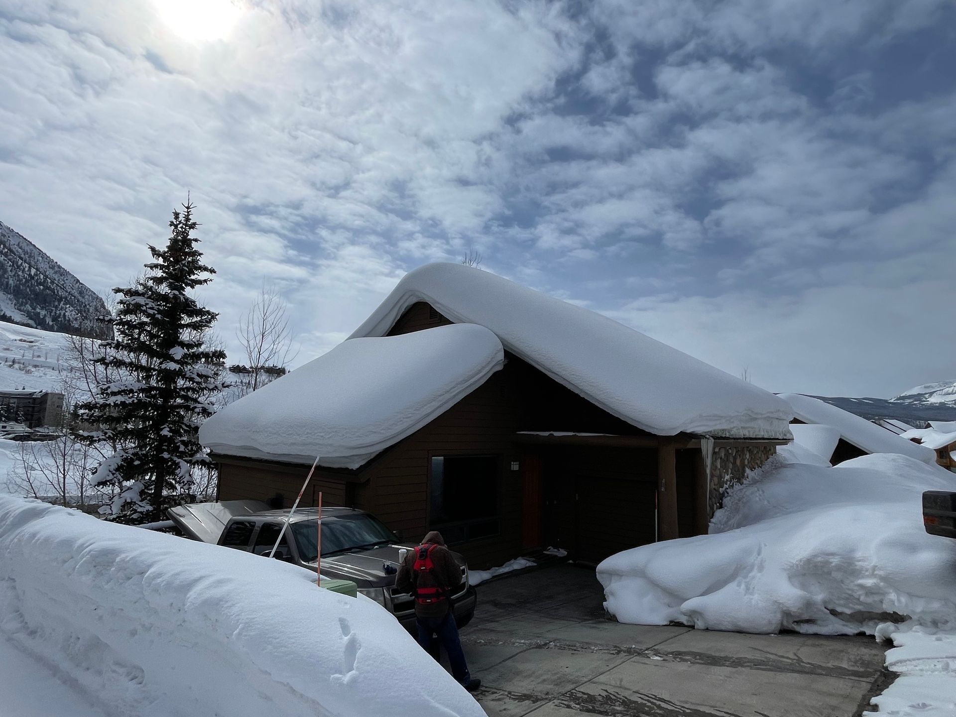 Snow-covered building in a mountainous setting, with a person walking towards a car partially buried in snow.