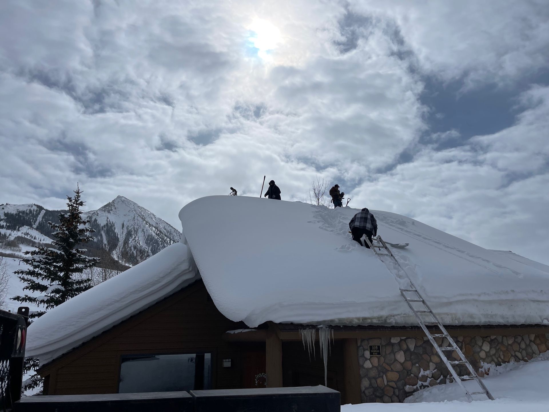 People clearing snow from a building's roof with ladders and shovels in a snowy mountain setting.
