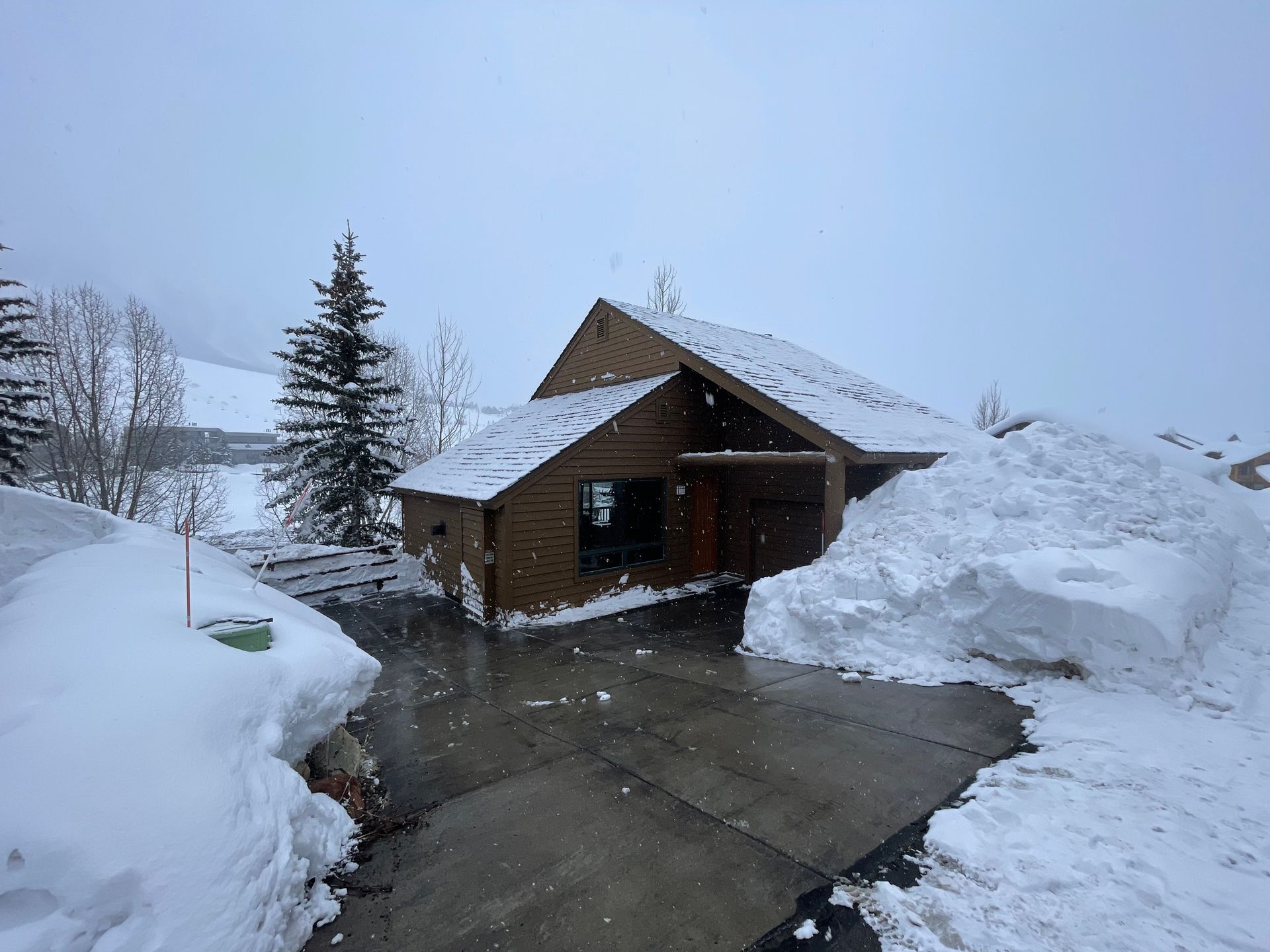 Snowy scene of a wooden house with a snow-covered roof, driveway, and large snowbanks.
