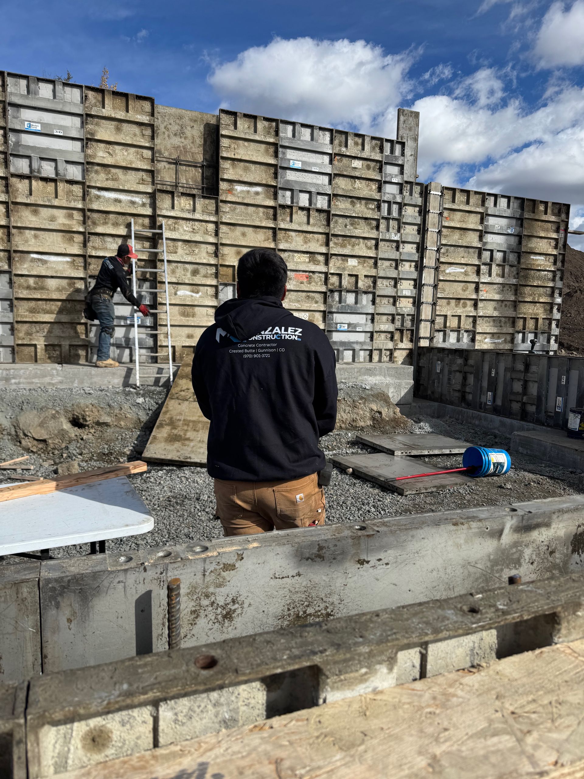 Construction site with workers, concrete forms, and equipment. Blue sky with clouds in the background.