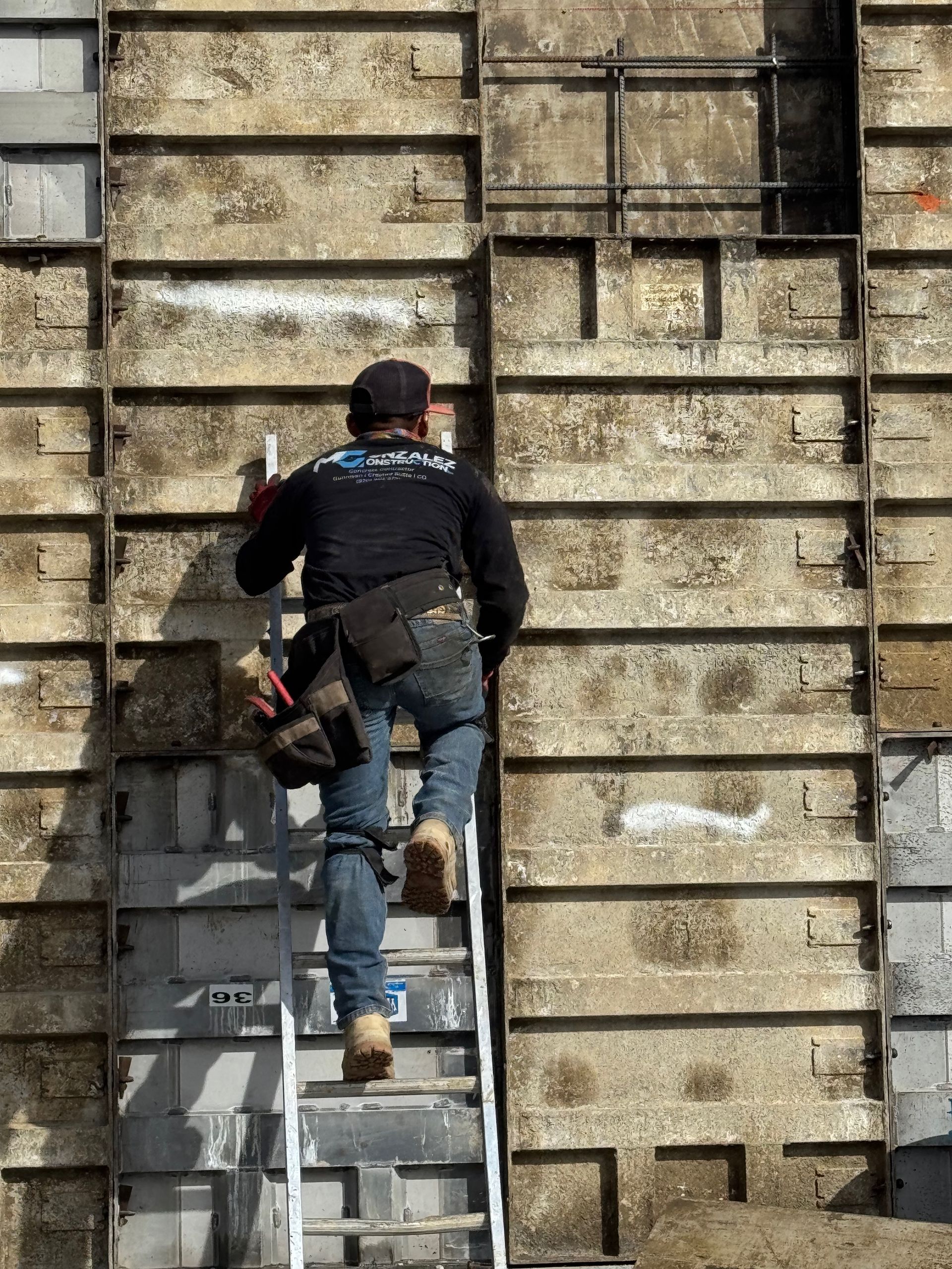 Construction worker climbs a ladder on a concrete wall, wearing work clothes and a tool belt.