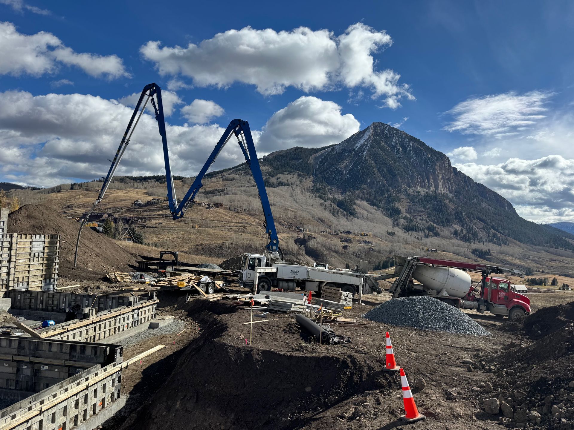 Construction site with concrete pump truck and a mountain in the background under a blue, cloudy sky.