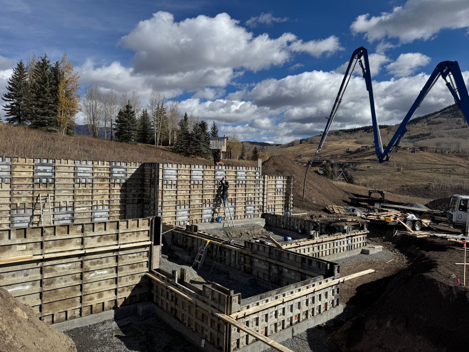 Construction site with concrete forms being filled; pump arm reaching, mountains and sky visible.