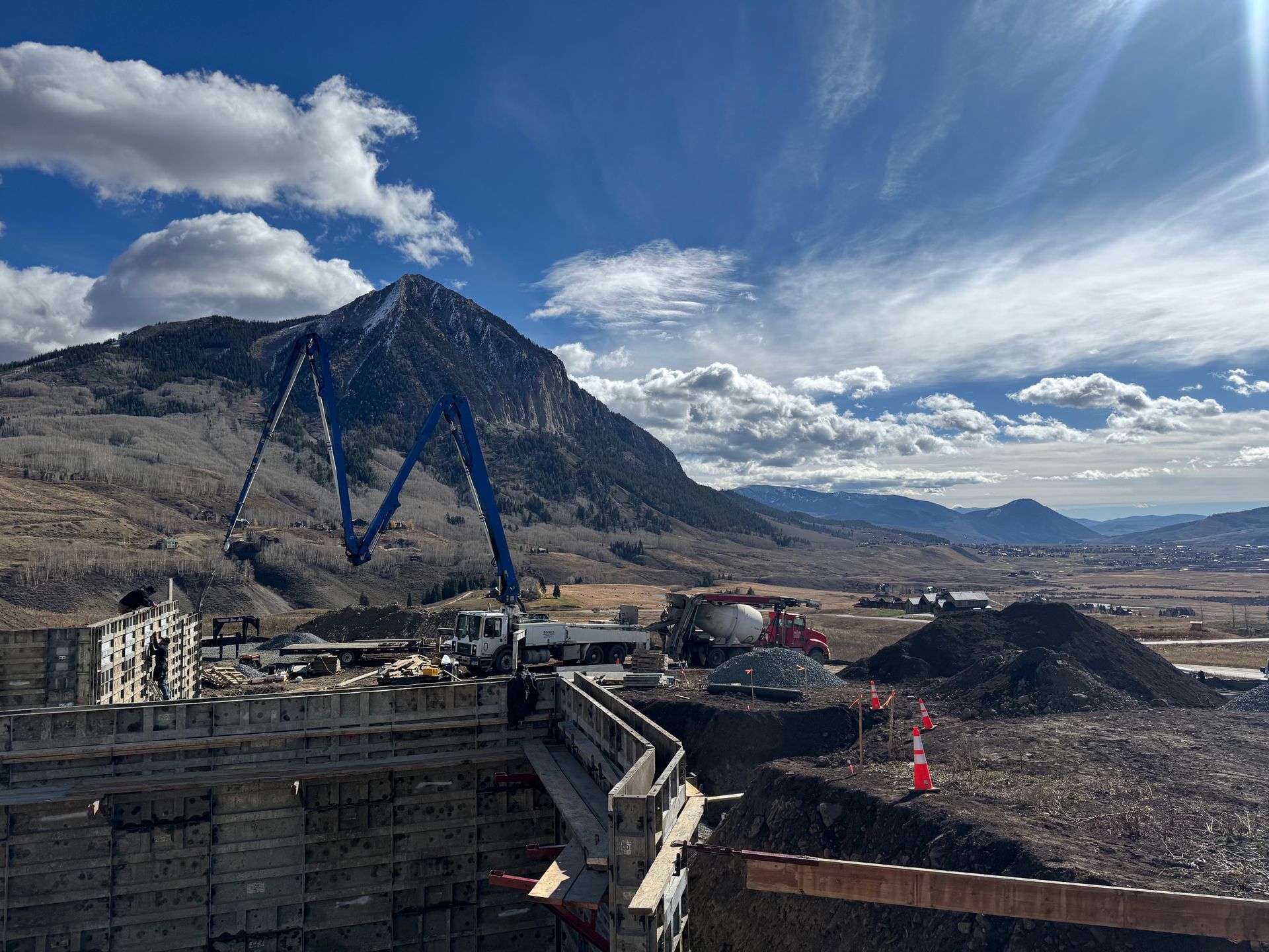 Construction site with a concrete pump truck, mountain in the background, under a blue sky with clouds.