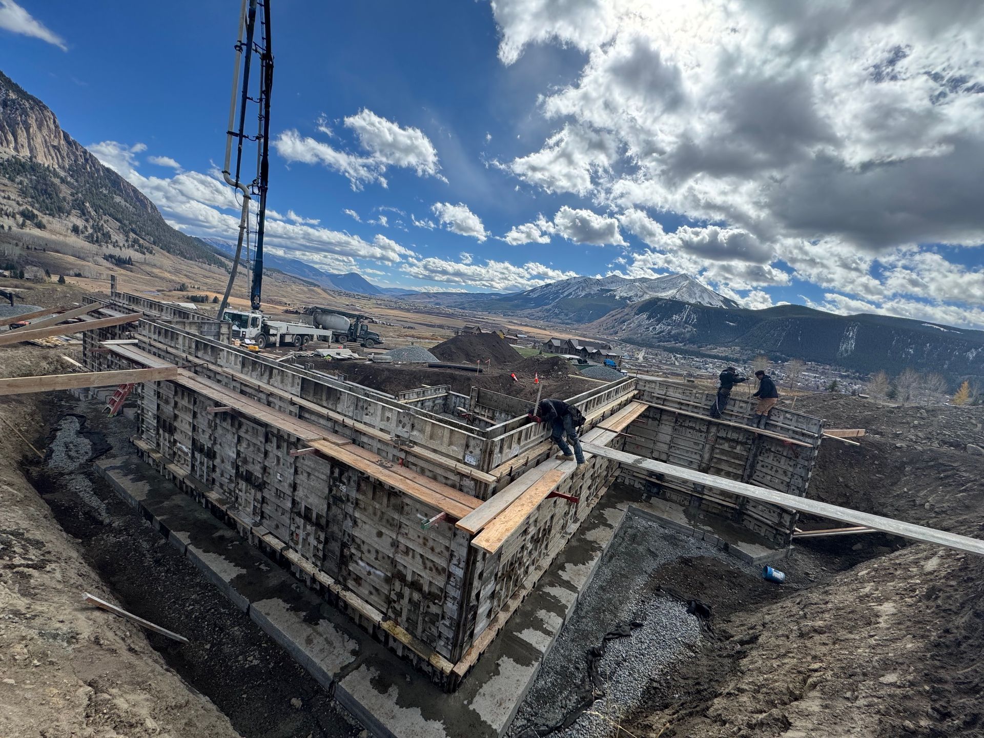 Construction site with concrete forms, workers, and a mountainous landscape under a cloudy sky.