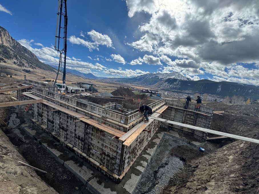 Concrete foundation construction with workers, pump truck, and mountain landscape under a partly cloudy sky.