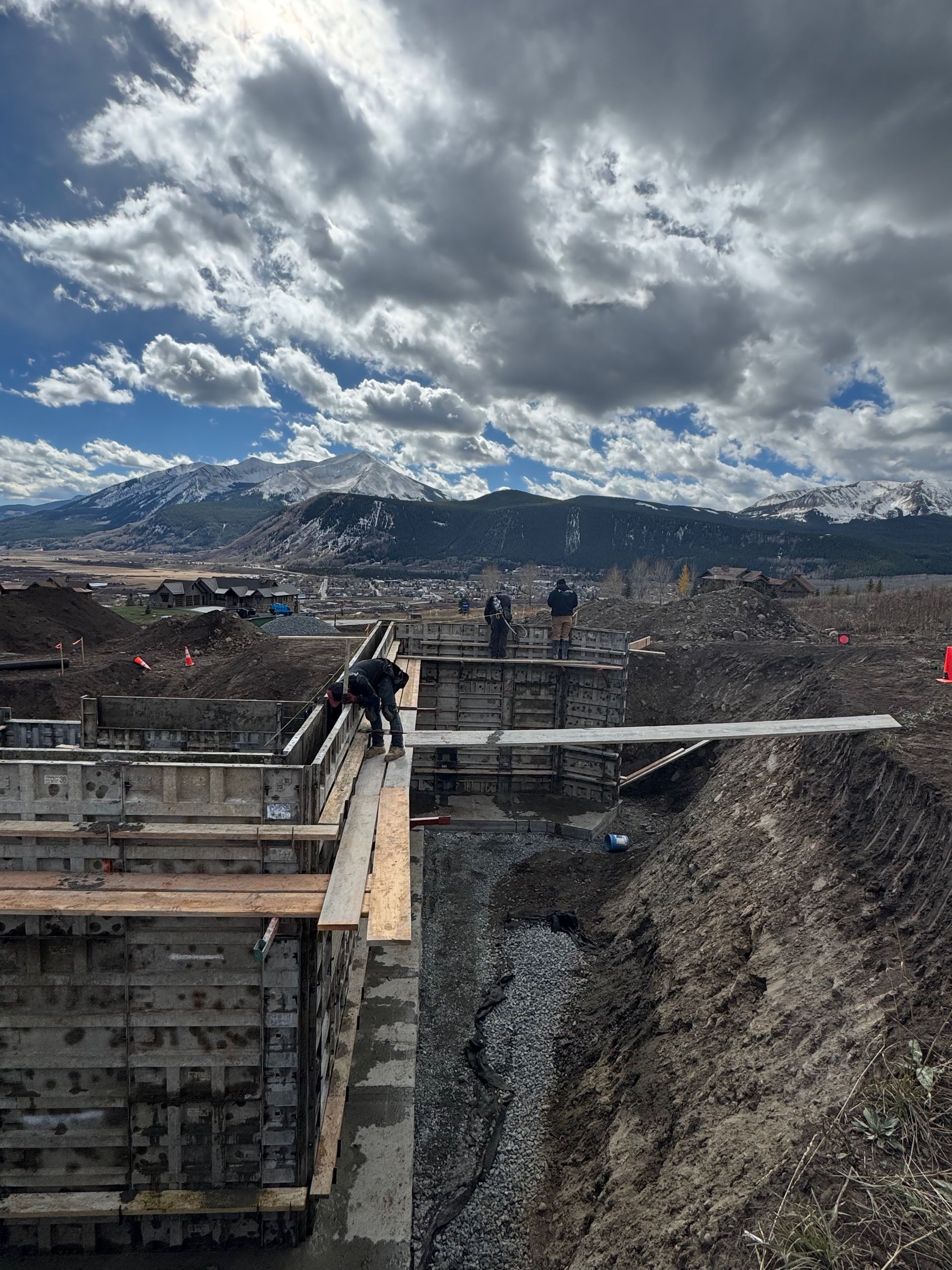 Construction workers building a structure outdoors with mountains in the background under a cloudy sky.