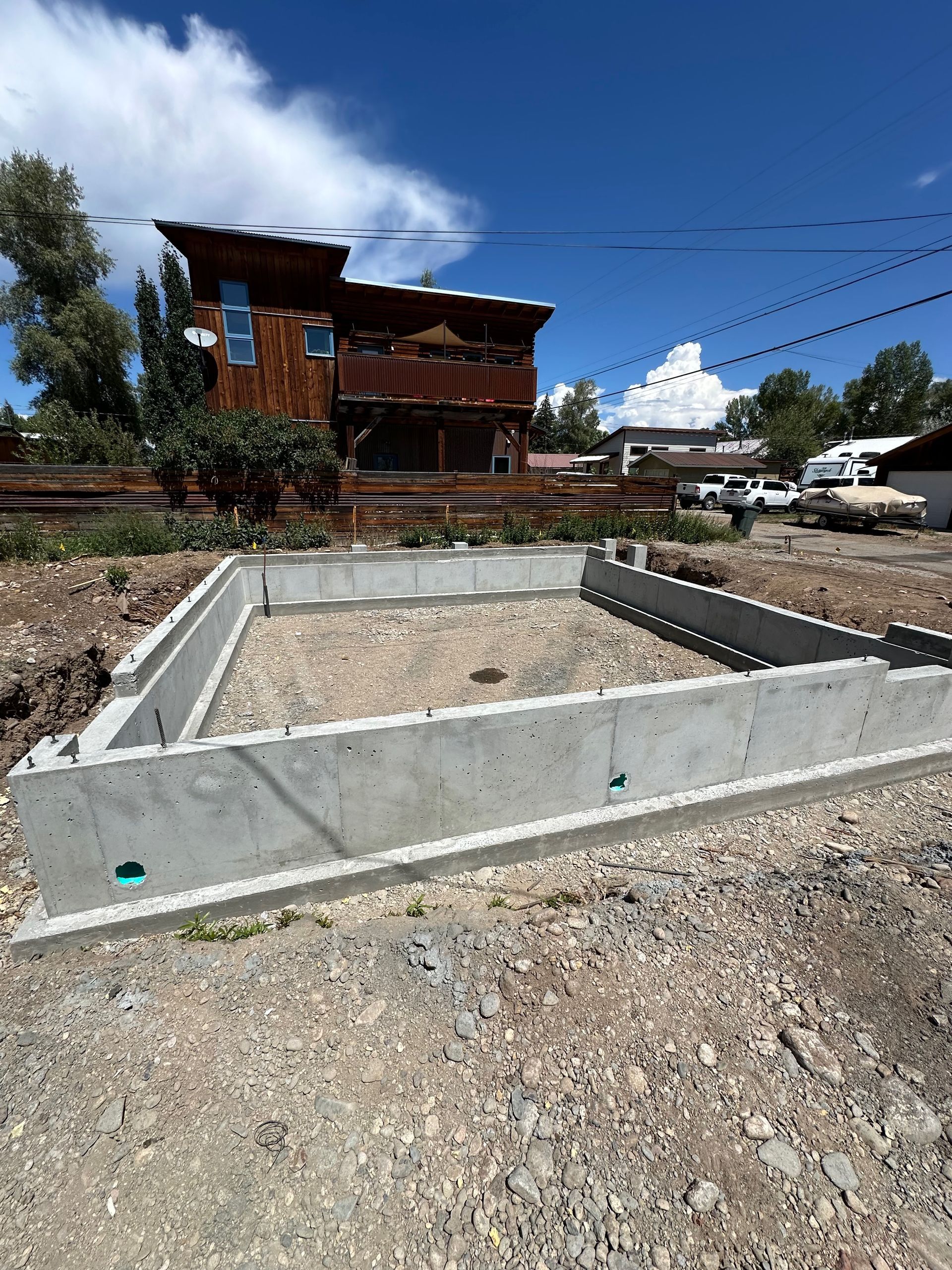 Concrete foundation for a building in a dirt lot, with a wooden house in the background on a sunny day.