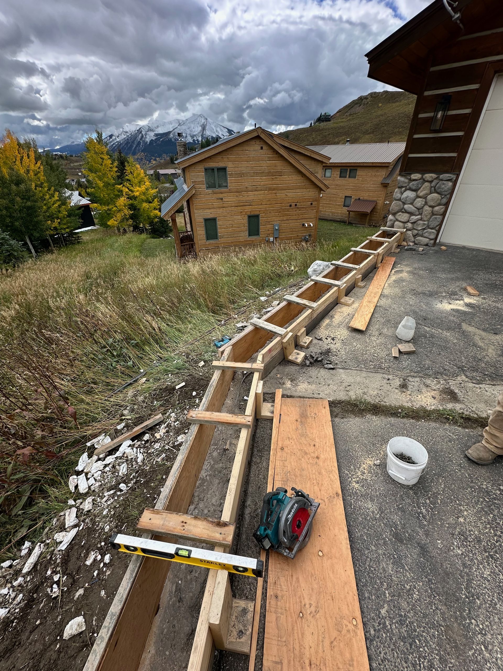 Construction site: Wooden forms for a wall being built outdoors, mountains and houses in the background.