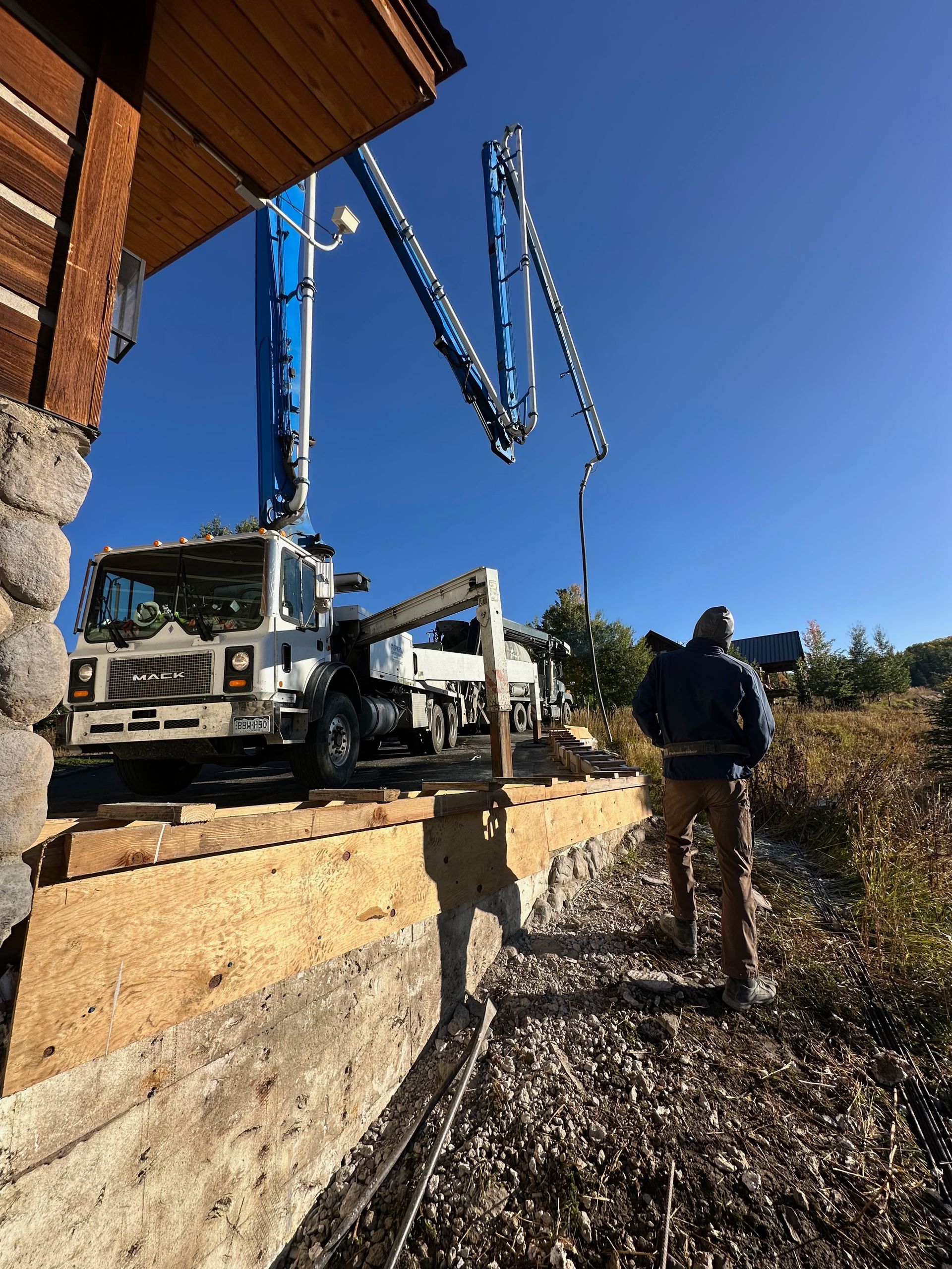 Concrete pump truck near a building; a person stands nearby. Blue sky overhead.