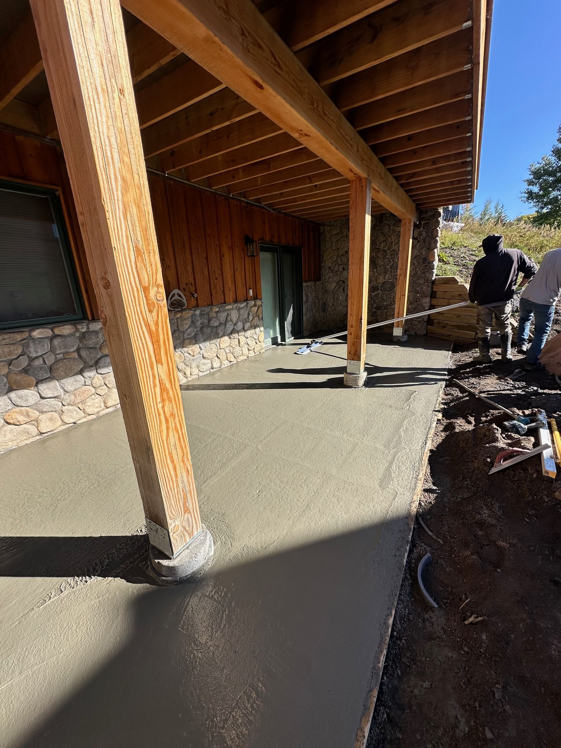Freshly poured concrete walkway under a wooden porch with workers in background.