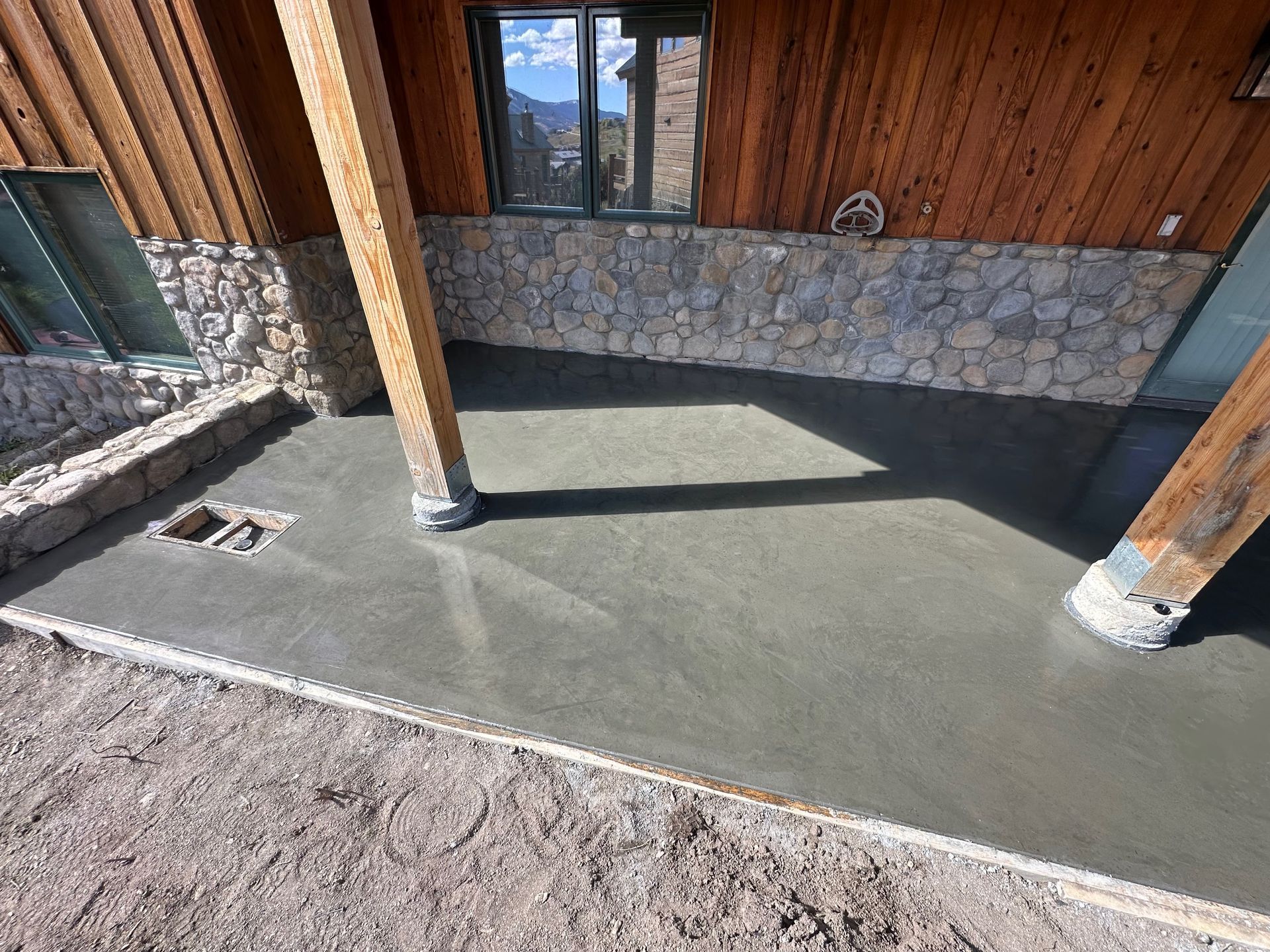 Freshly poured concrete patio under a wooden overhang, next to a stone wall.