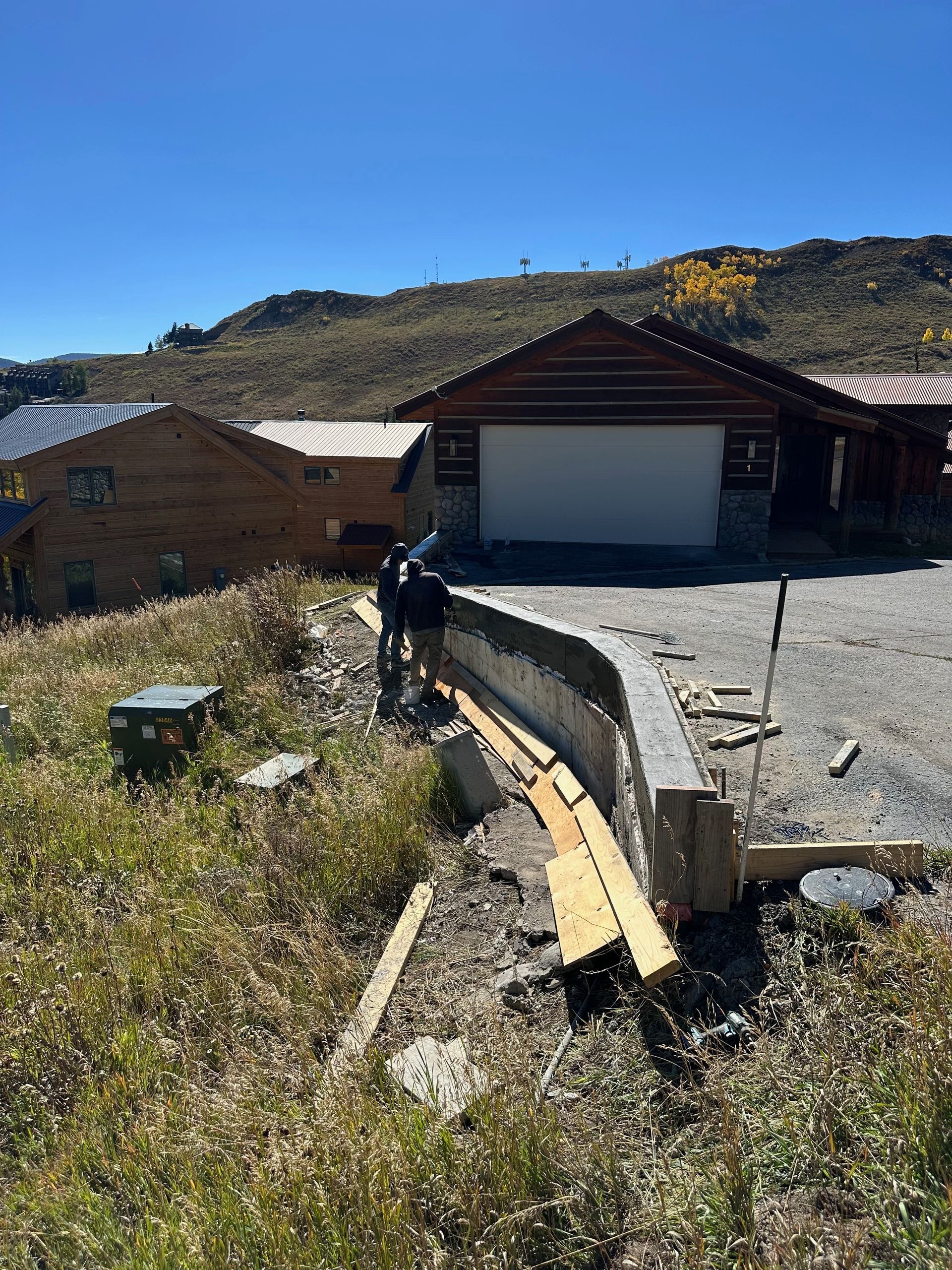 Two people working on concrete structure in front of a rustic garage, surrounded by dry grass and a mountain landscape.