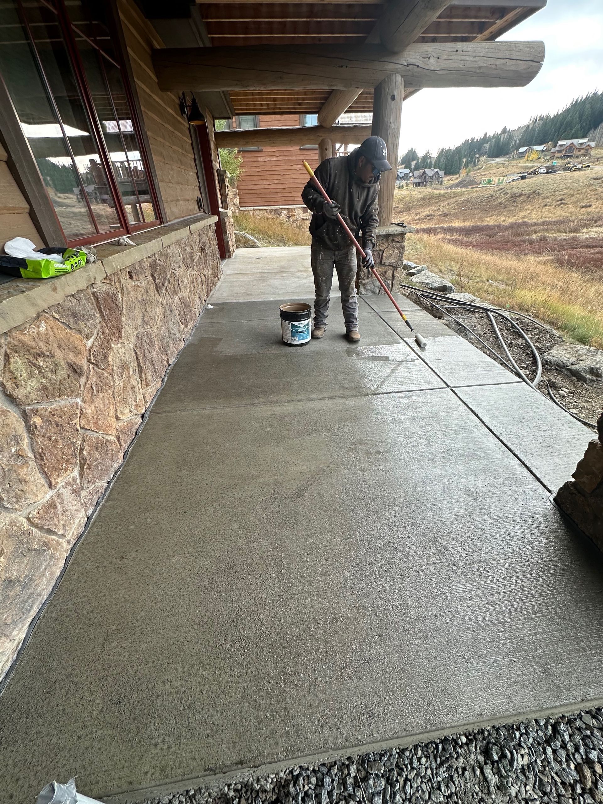 Person applying sealant to a concrete walkway next to a stone wall. Bucket and equipment visible.