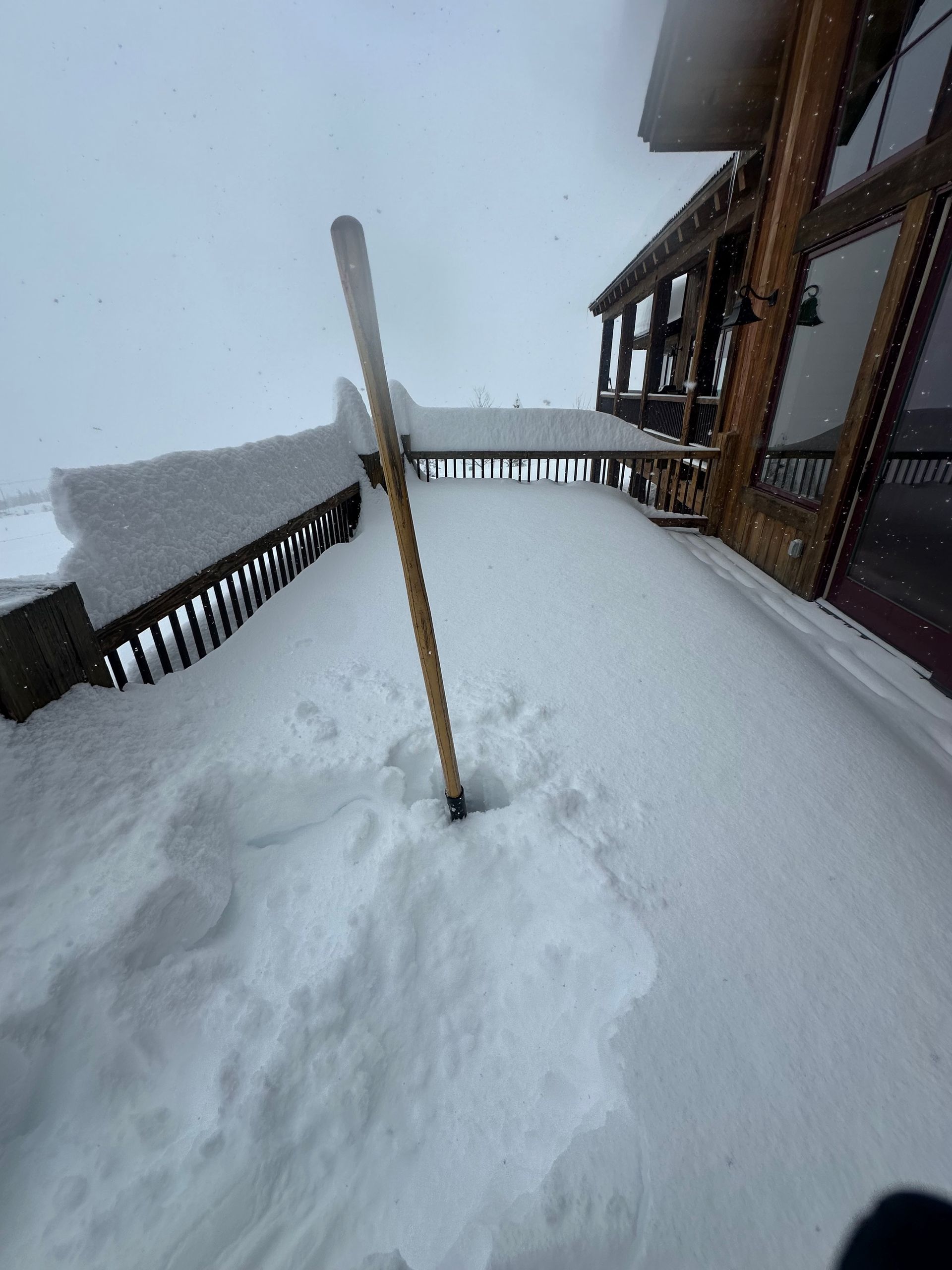 Snow-covered wooden deck with shovel. Snowfall visible; adjacent to a building with windows.