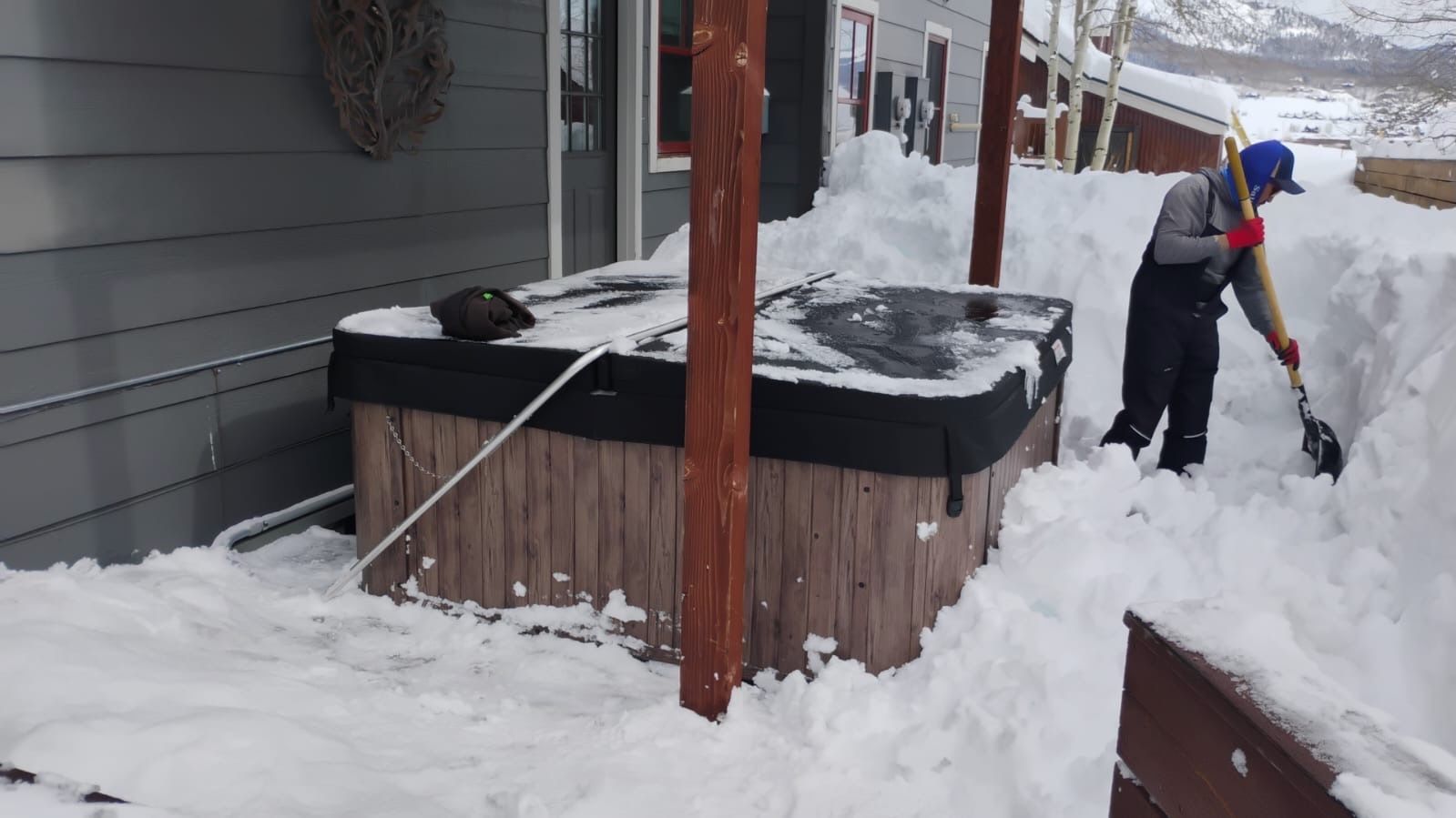 Person shoveling snow from around a hot tub on a snowy deck, near a house.