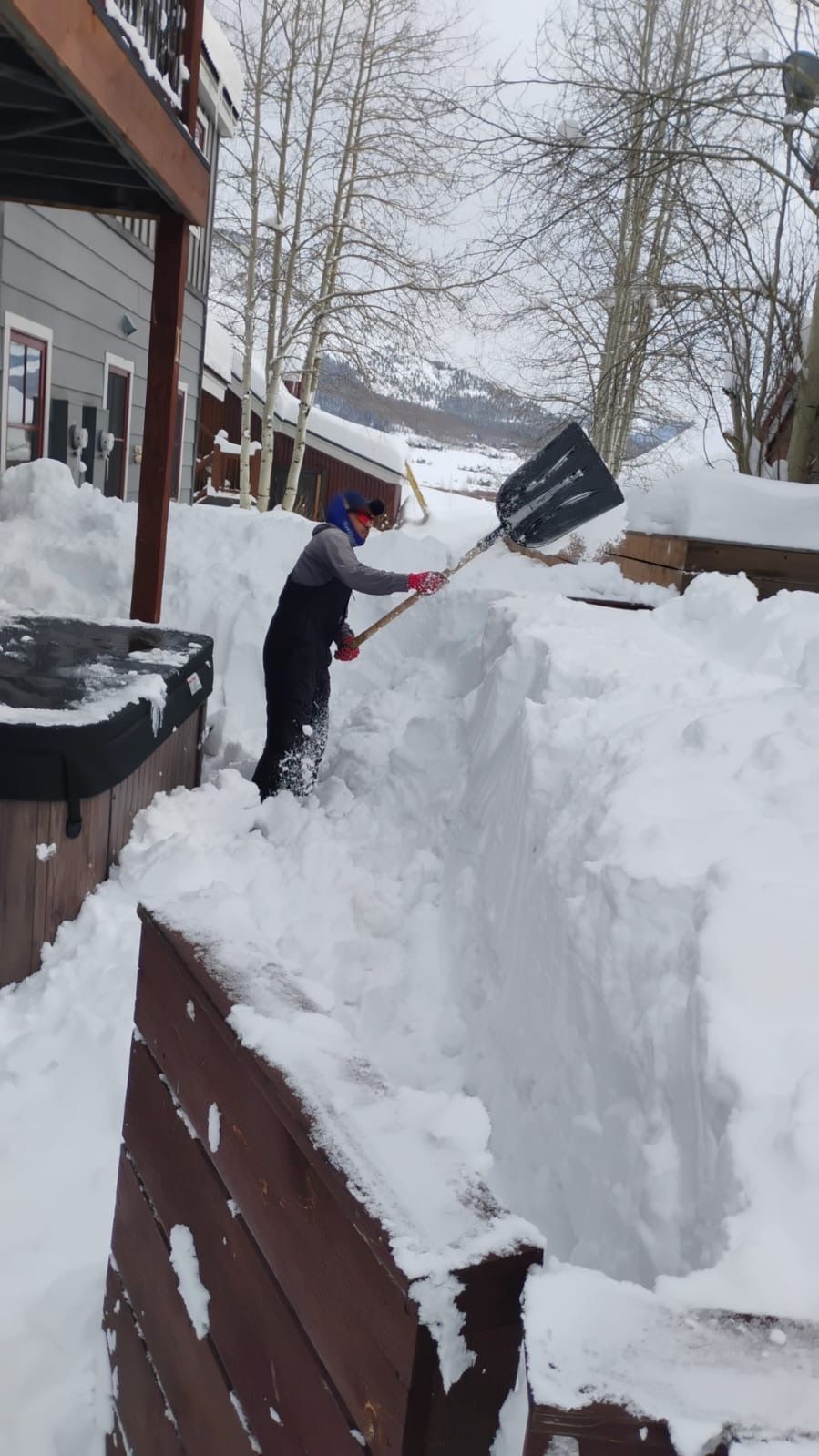 Person shoveling snow next to a hot tub. Snowy winter setting with a wooden deck and buildings.
