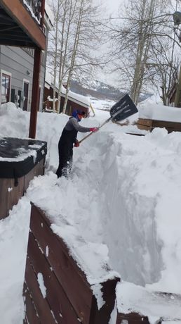 Person shoveling snow from a narrow walkway next to a hot tub and wooden deck in a snowy, outdoor setting.