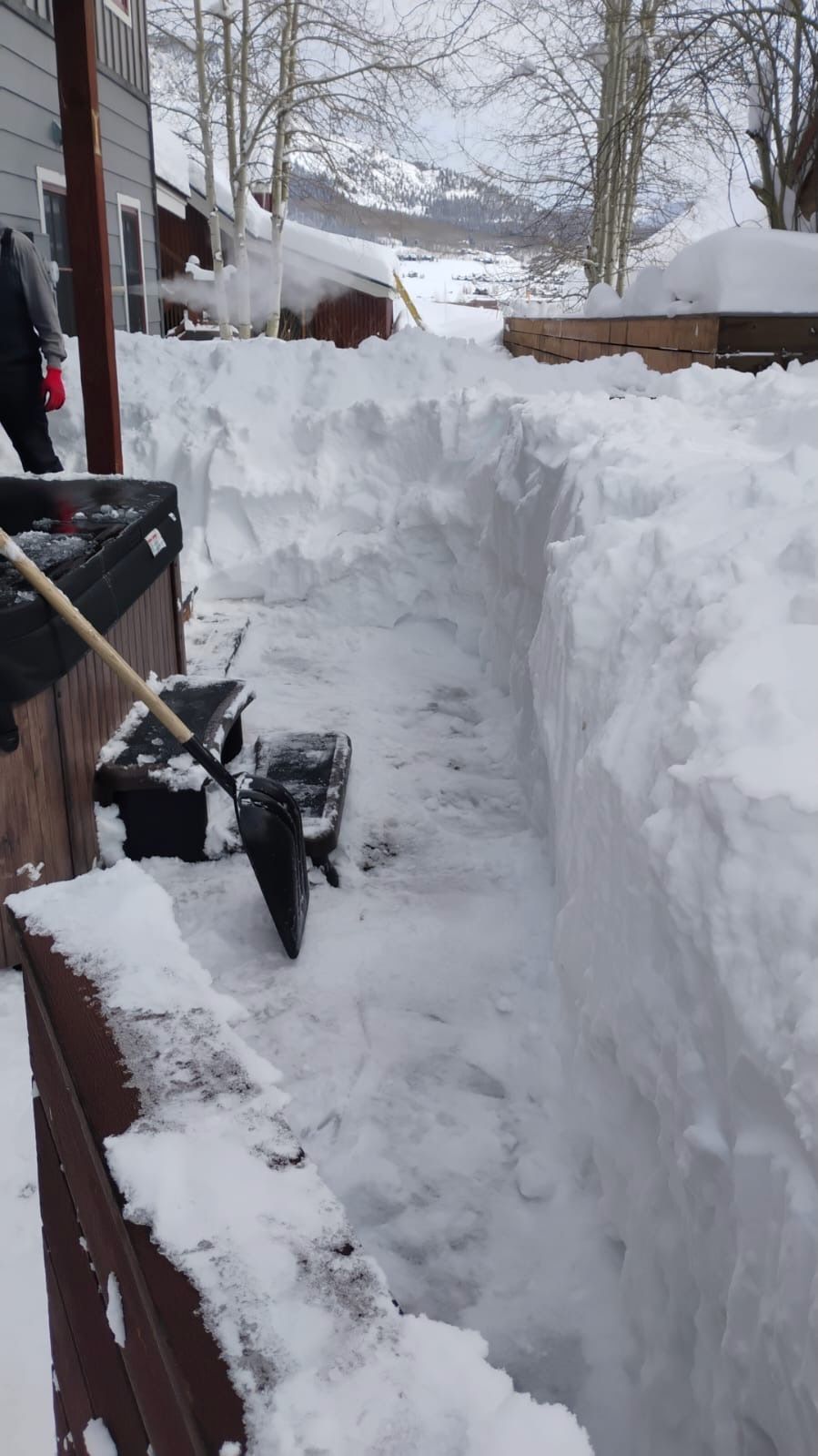 Person shoveling a path through deep snow on a deck. Snow walls are visible with a mountainous backdrop.