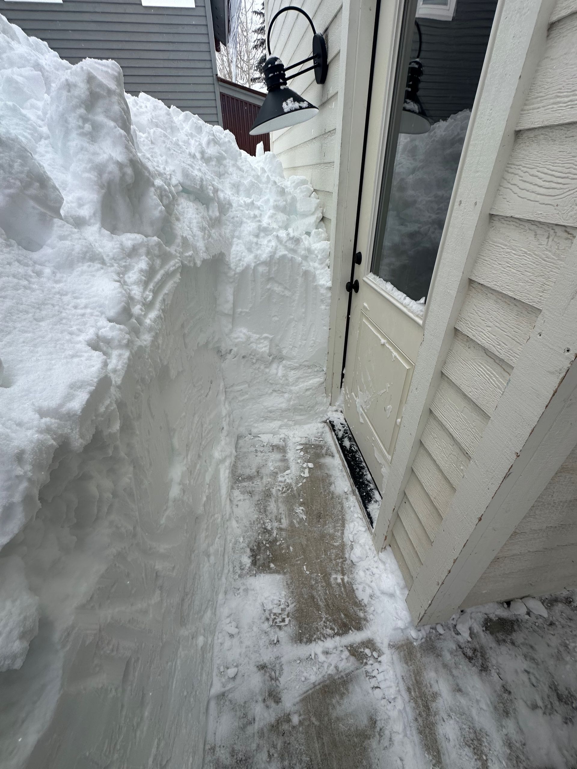 A snowy pathway, cleared to a door, is flanked by a large snowdrift against a white building.