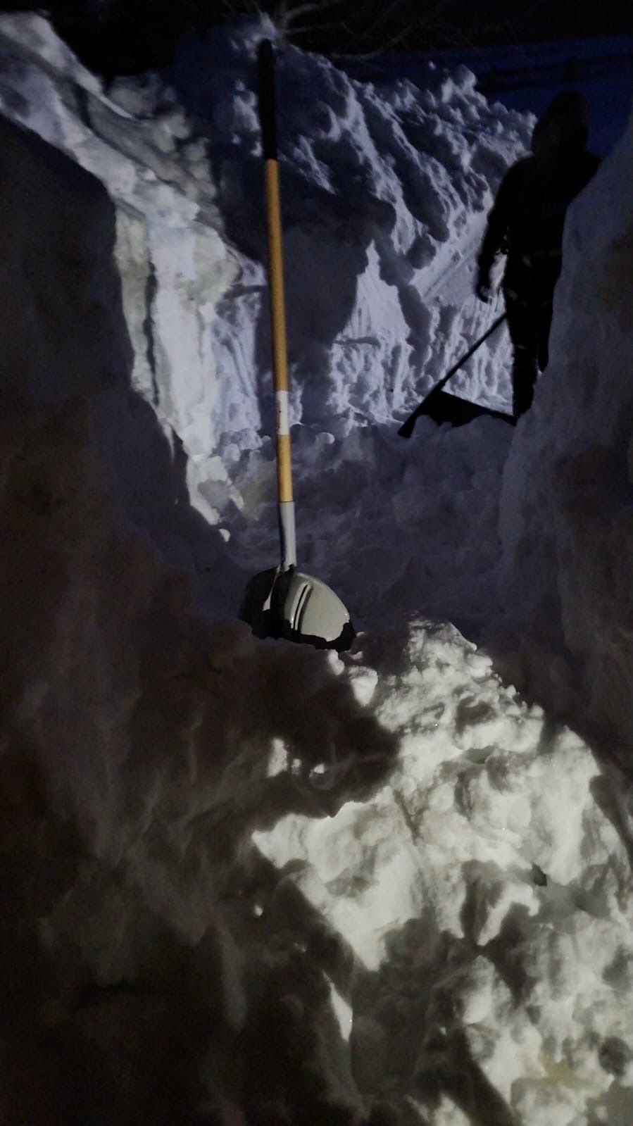 Person digging in deep snow, illuminated by light. A shovel rests in the snow, dark night setting.
