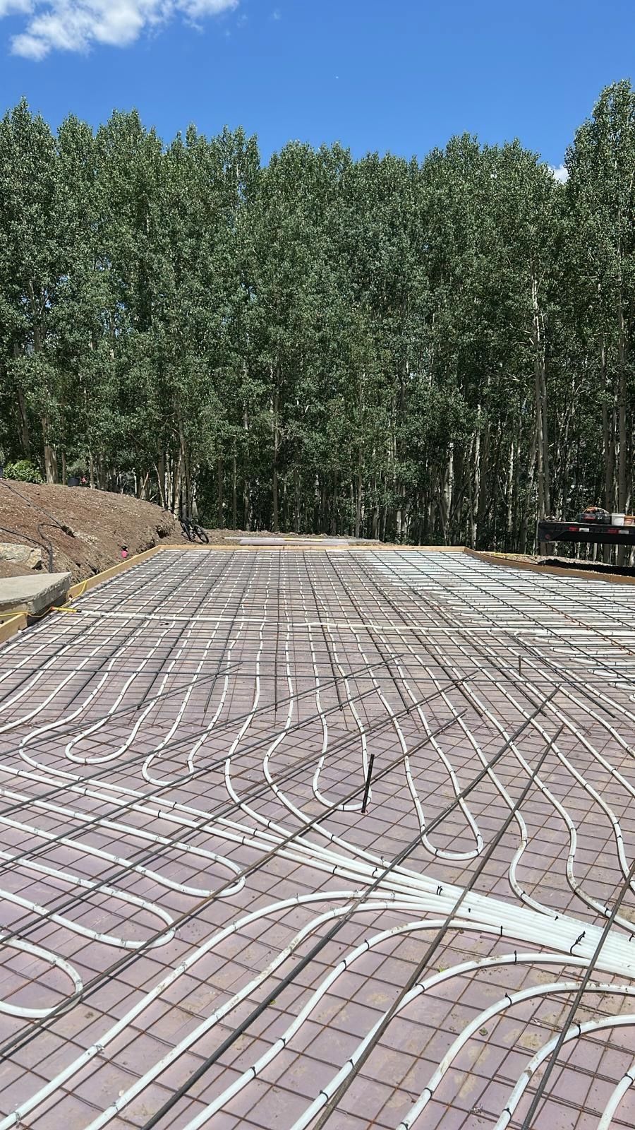 Pipes laid in wavy patterns across a concrete base, grid of metal bars. Trees in the background, blue sky.
