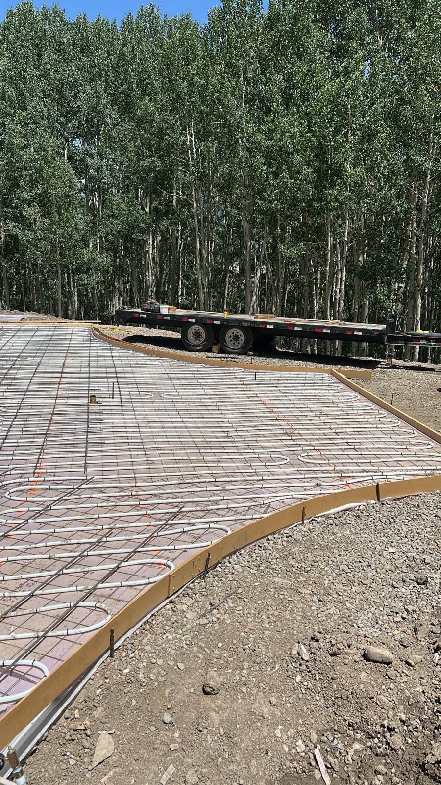 Construction site with a concrete form and rebar. A flatbed trailer sits in the background near trees.
