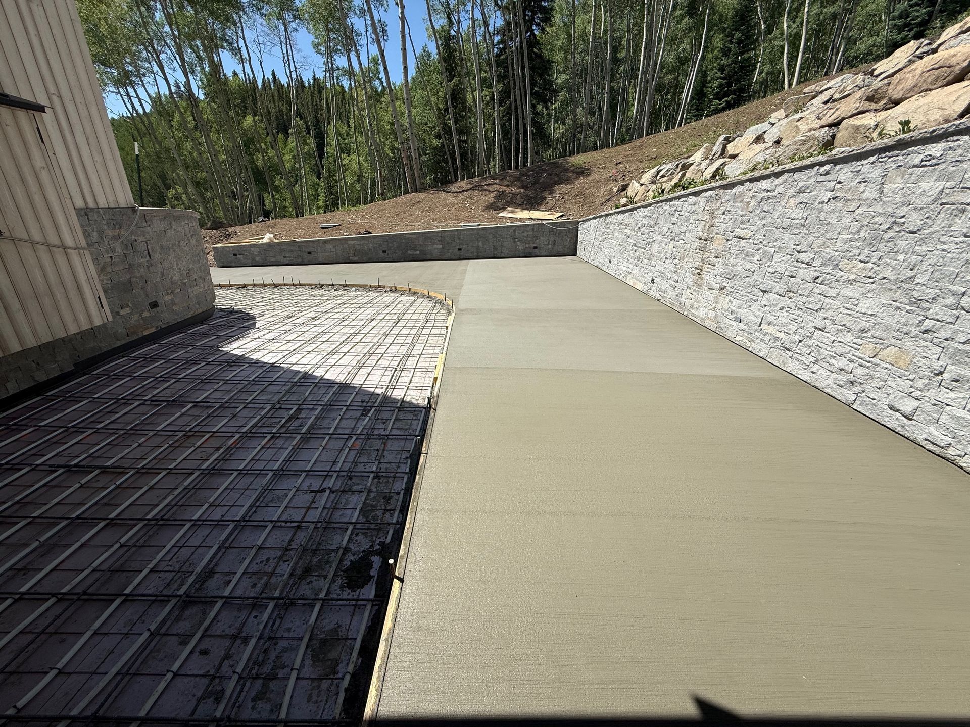 Freshly poured concrete pathway next to a rebar-covered area, set against a retaining wall and trees.