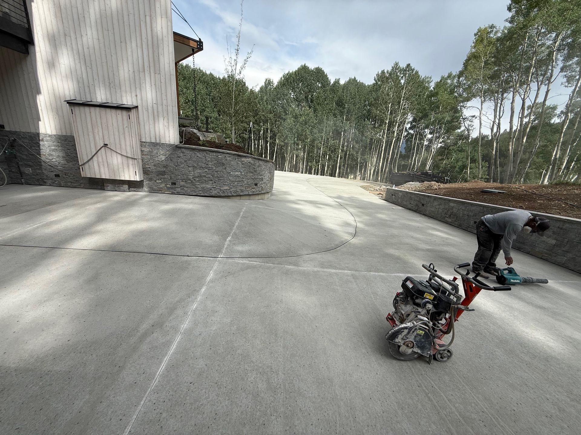 Person using a saw to cut concrete on a driveway, next to a concrete vibrator machine.