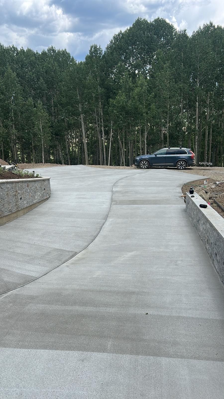 Concrete driveway curving uphill, with a car parked in front of a forest of trees, gray stone walls on either side.