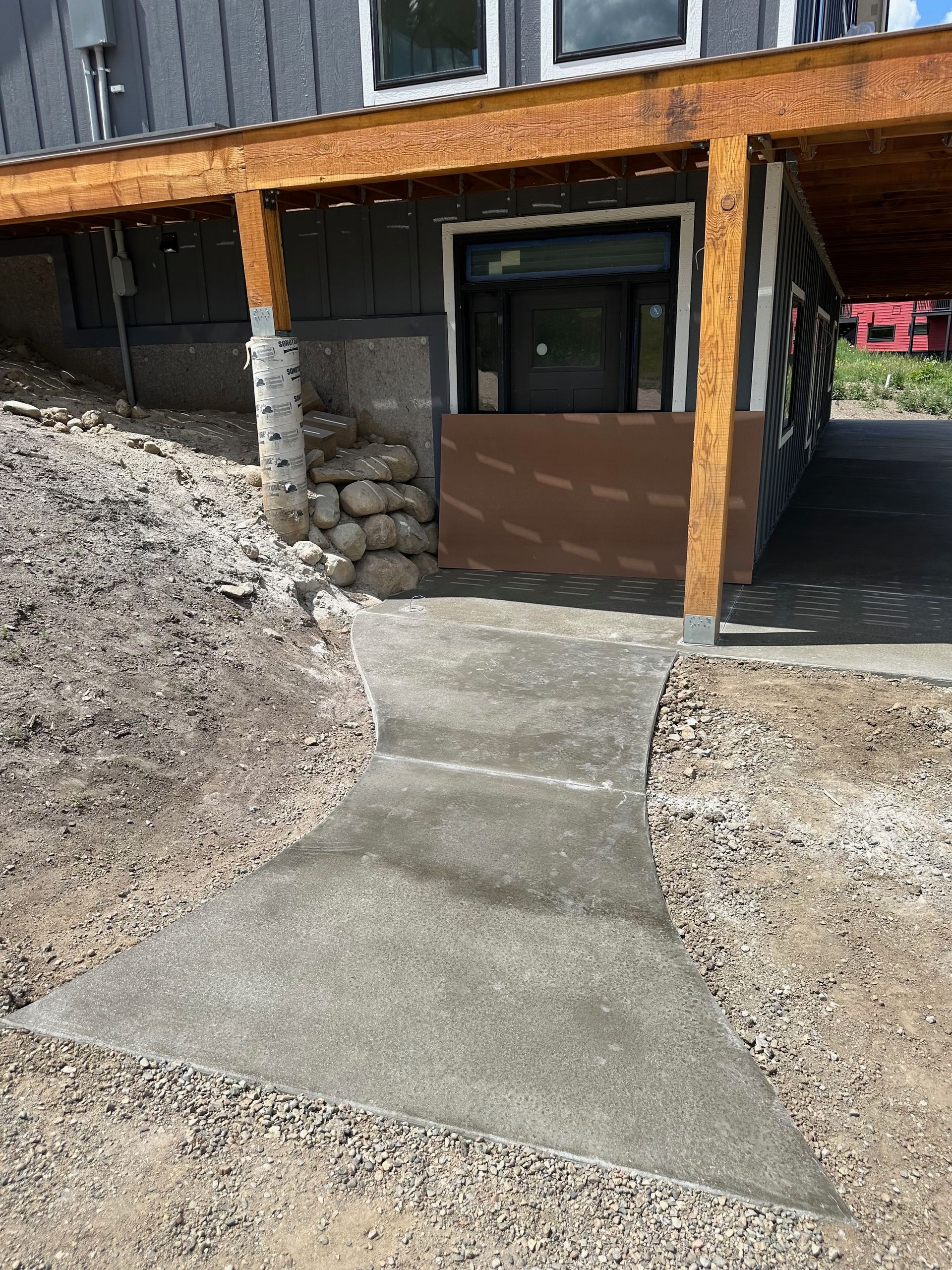 Concrete walkway leading to a building with a covered patio. Gray siding and wooden beams are visible.