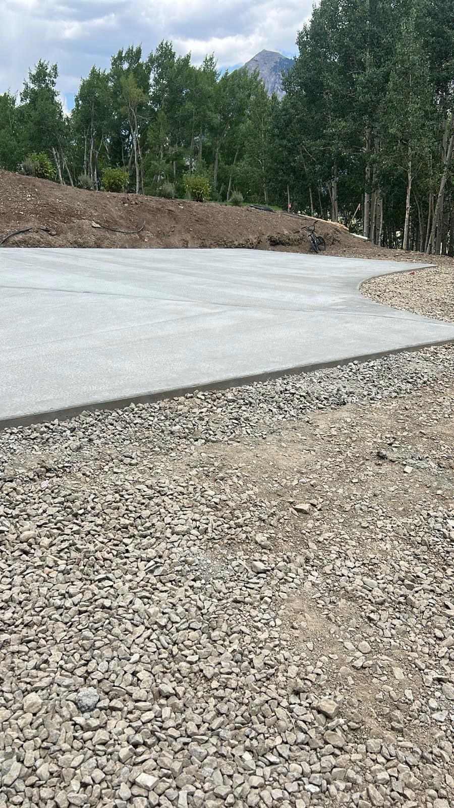 Concrete pad next to gravel, with dirt hill and trees in the background.