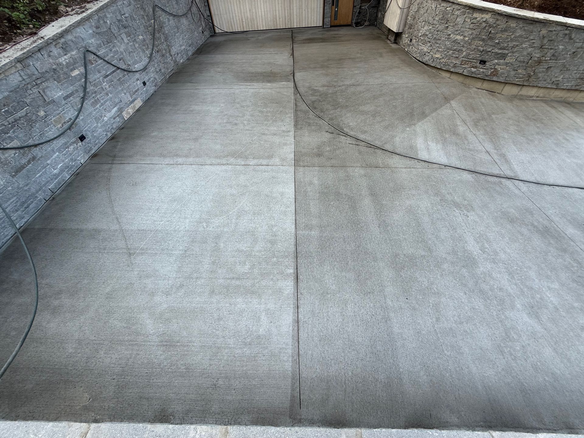 Concrete driveway, half cleaned, showing a visible contrast. Gray concrete, surrounded by stone walls.