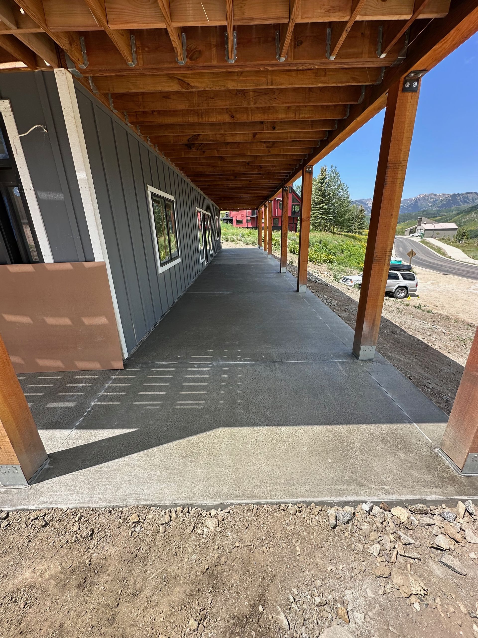 Covered concrete walkway outside a building with gray siding, wooden beams, and a gravel driveway.