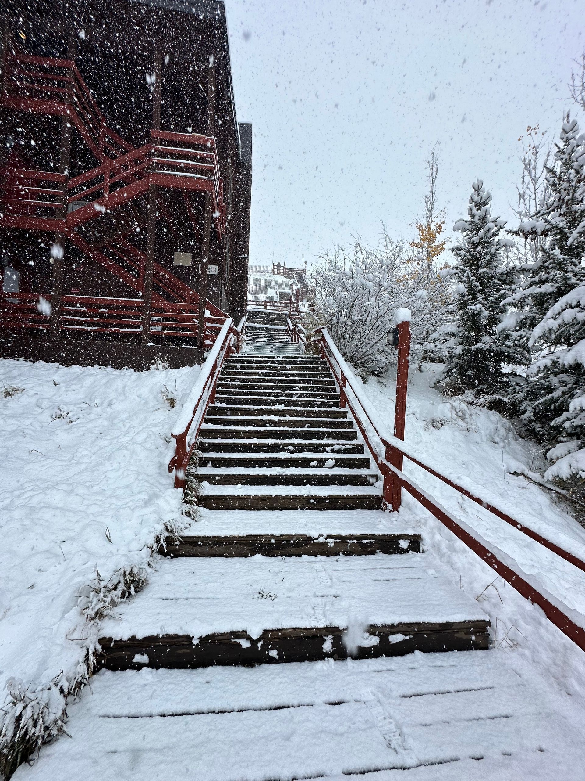 Snowy outdoor staircase leading up to a red-sided building, with snow-covered trees and railing.