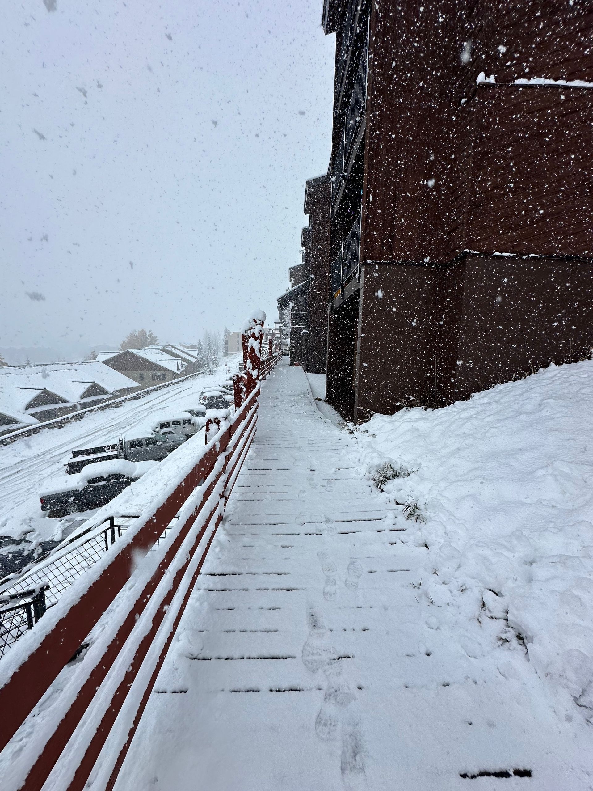 Snowy walkway next to a red-railed building and a parking lot, with falling snow.