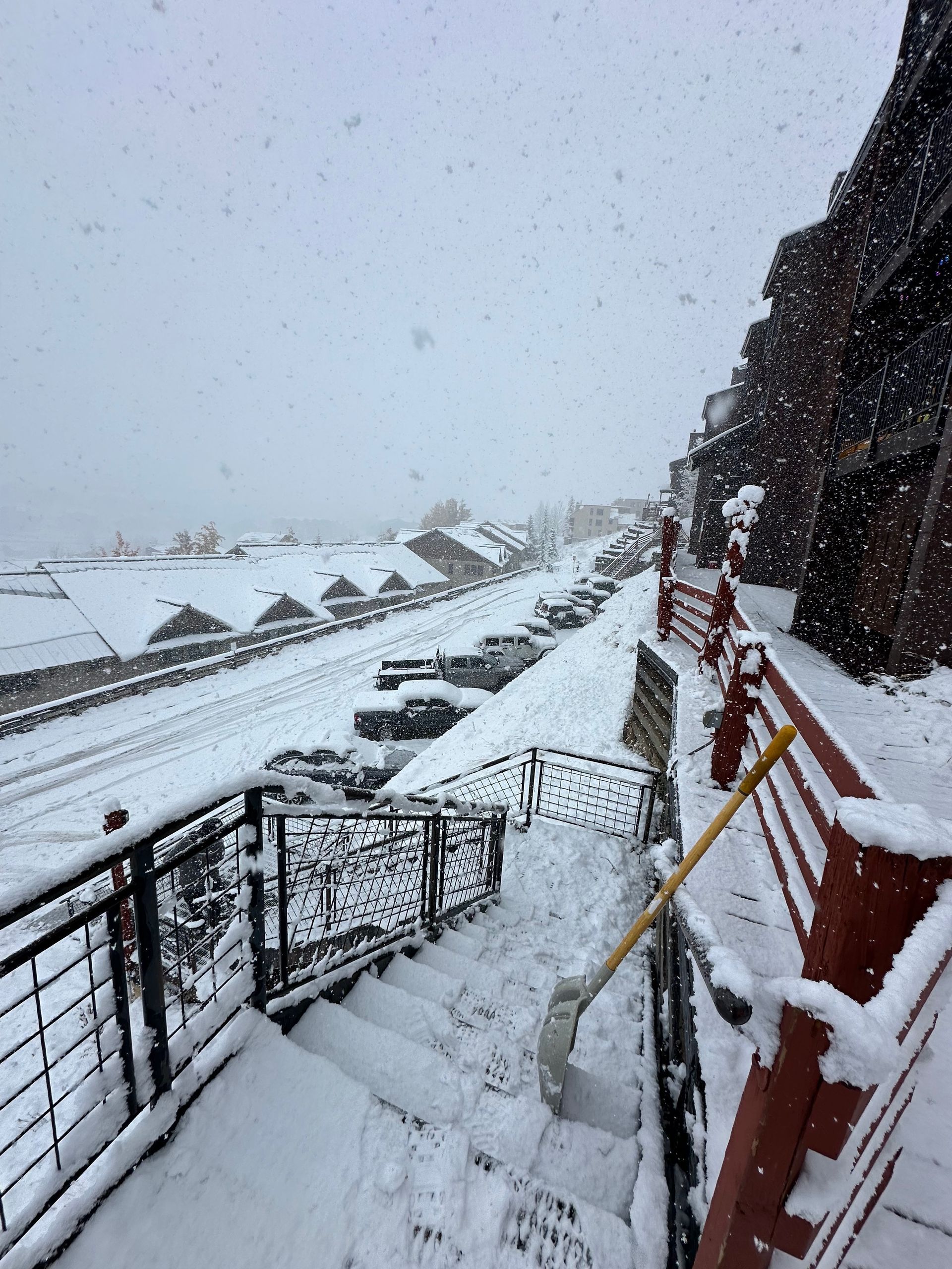 Snow-covered outdoor stairs with a shovel, leading to a street with cars and buildings in a snowy landscape.