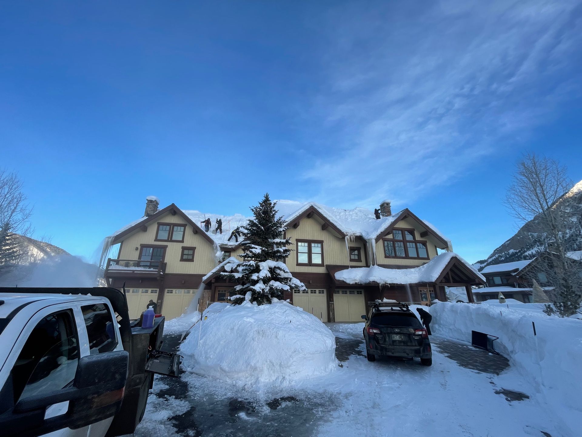Snow-covered house with driveway and vehicles. A person shovels snow. Blue sky and snow-covered trees in the background.