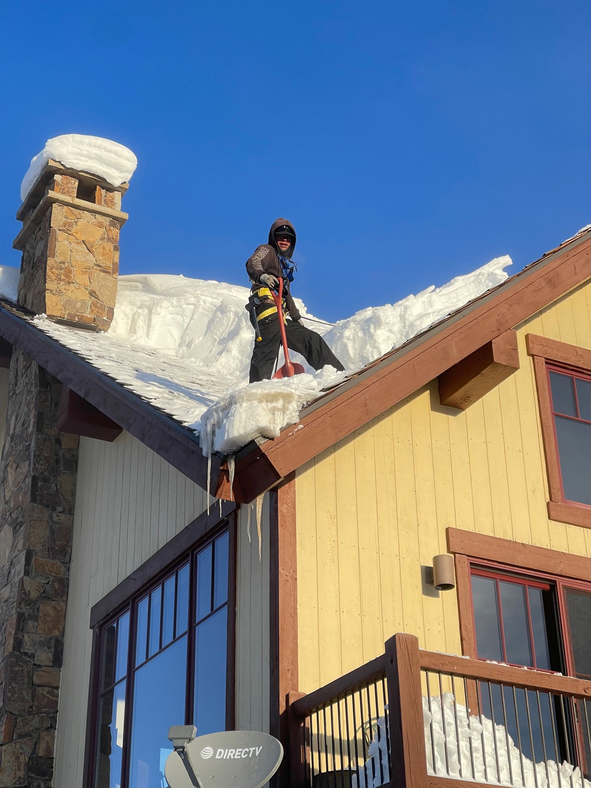 Person on a snow-covered roof clearing snow. Sunny day, yellow house with stone chimney.