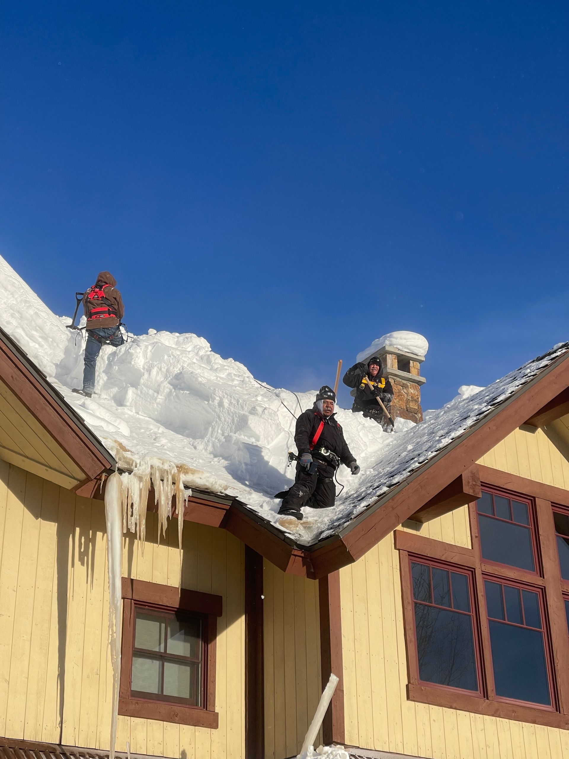 Three people shoveling snow from a yellow house roof under a clear blue sky.