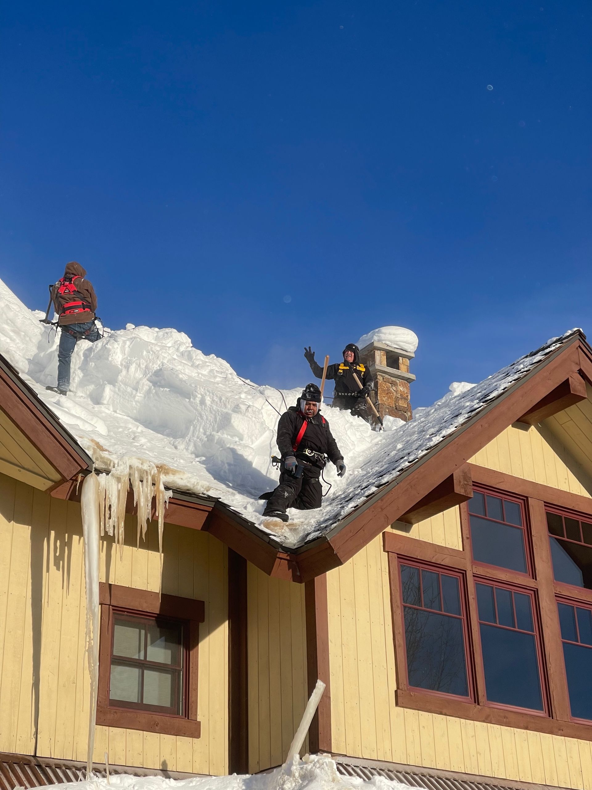 People clearing snow from a roof.  Yellow house, snowy roof, clear blue sky, icicles.