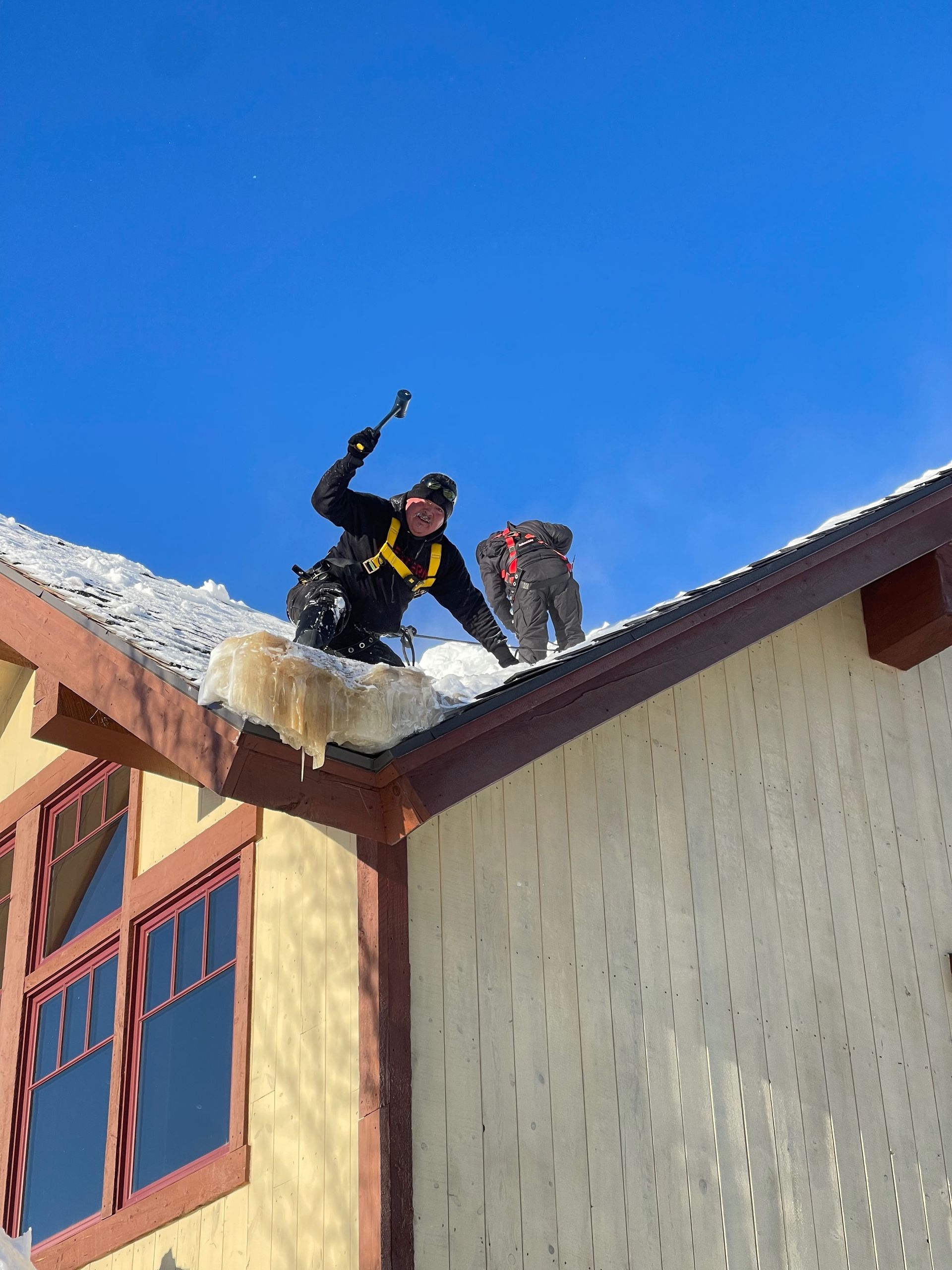Two people on a snow-covered roof clearing ice. One swings a tool; clear blue sky.