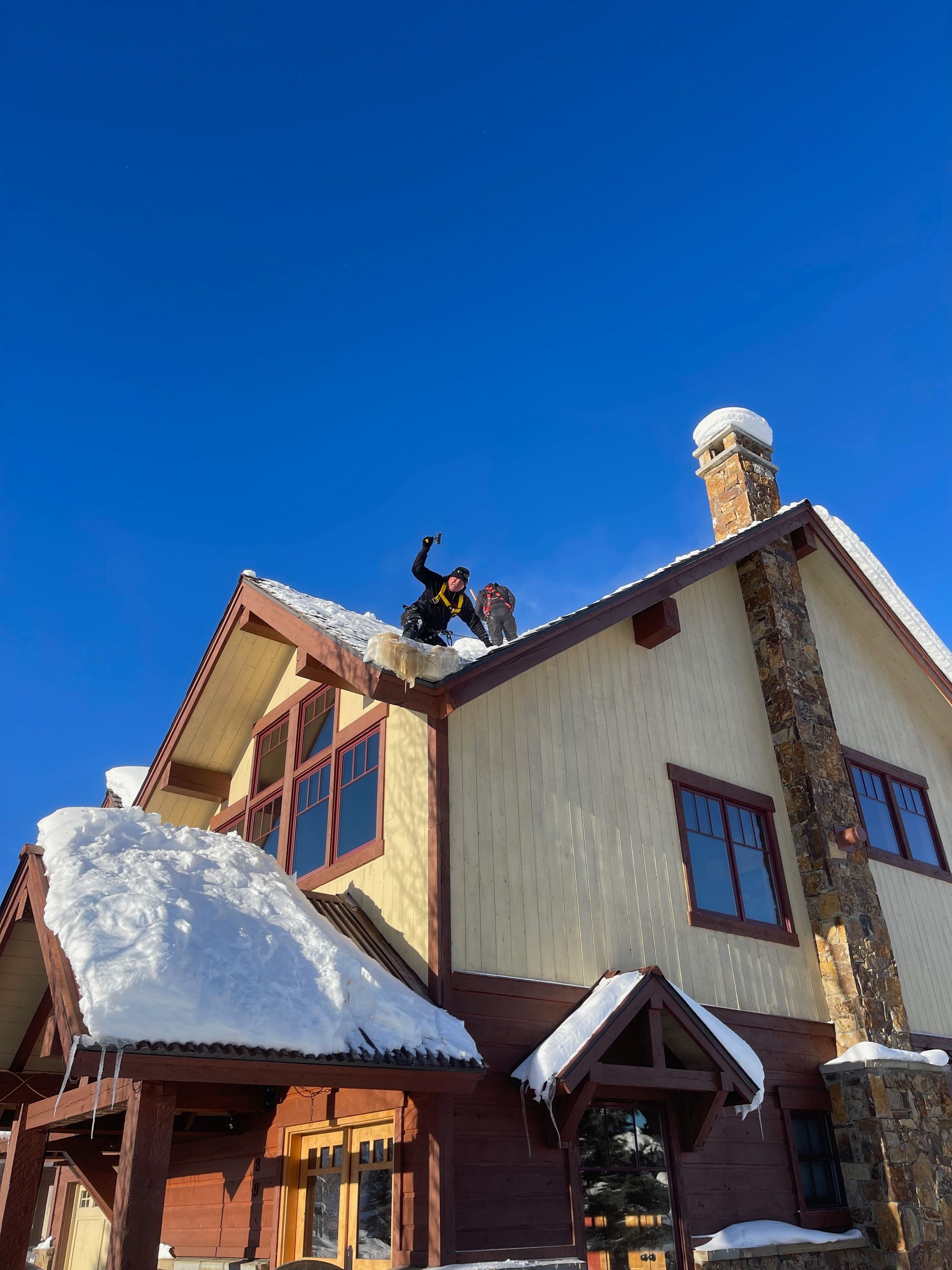 Person on snow-covered roof, clearing snow with a tool, against a bright blue sky.