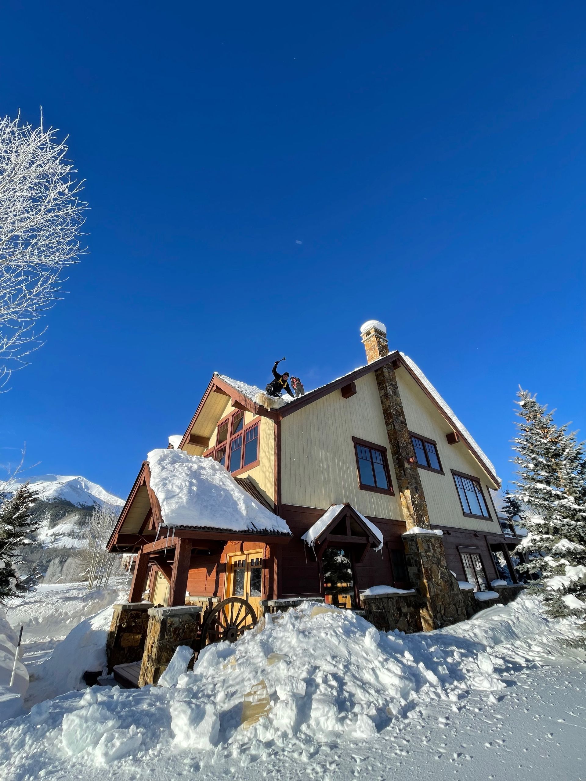 Snow-covered house with stone chimney, set against a bright blue winter sky. Snow falling from the roof.