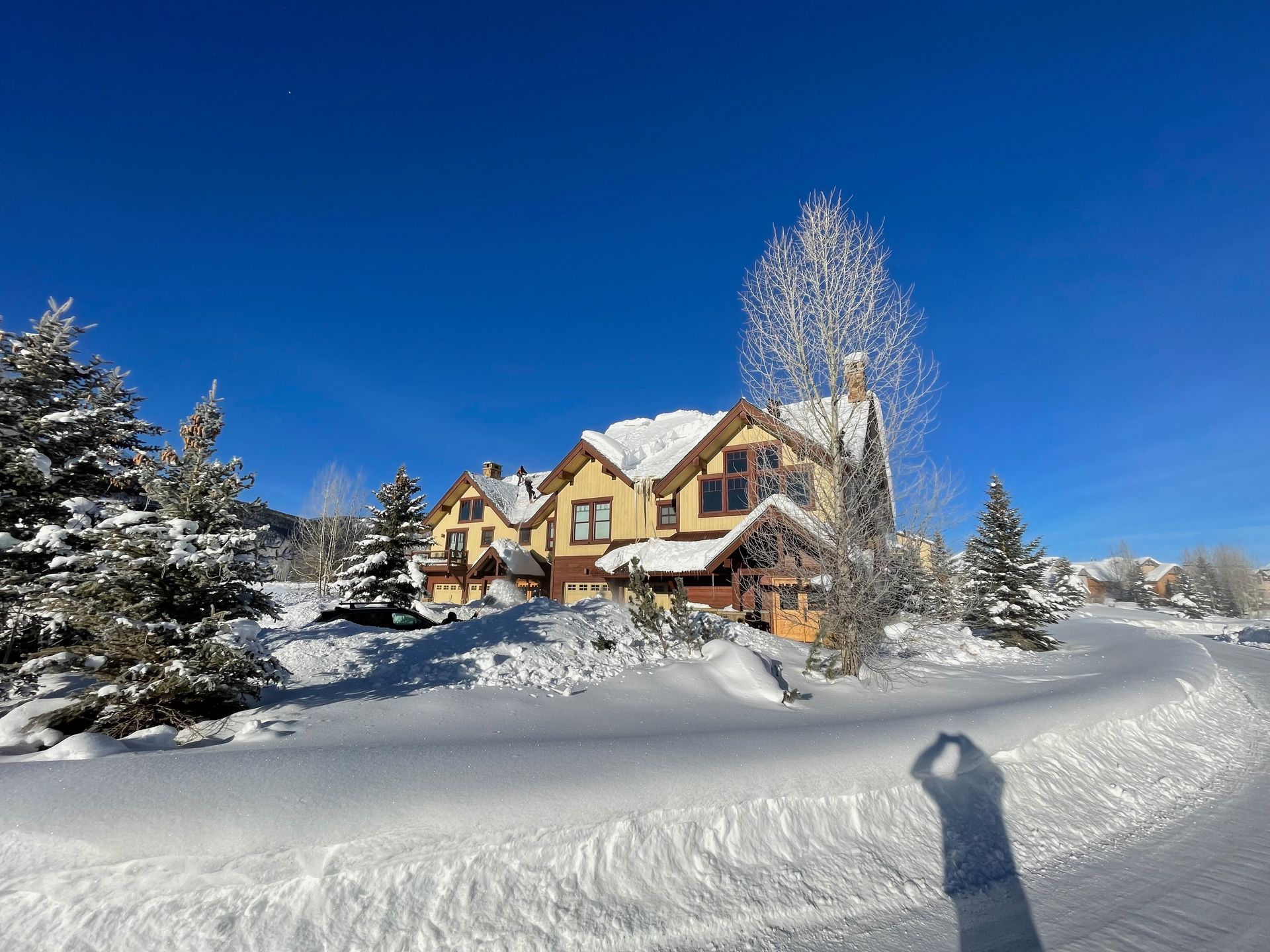 Snow-covered house with yellow siding and a snowy road under a bright blue sky.