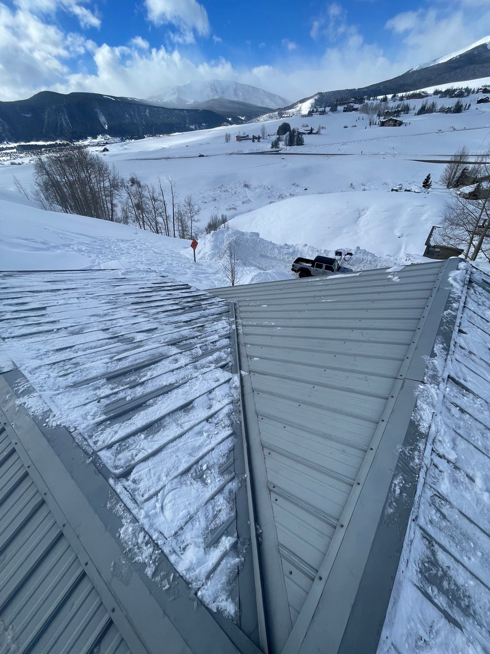 Snow-covered metal roof in the foreground, with a snowy mountain landscape in the background under a blue sky.