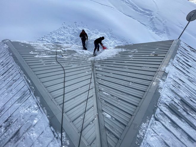 Two people shoveling snow from a metal roof in a snowy mountain setting.